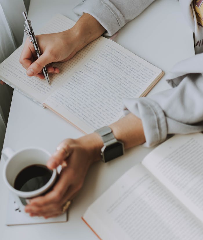 Close-up of a person writing in a notebook with coffee on a desk. Ideal for workplace or study themes.